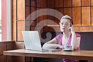 Beautiful thinkful young girl freelancer with short hair, in pink t-shirt and eyeglasses is sitting in cafe and working on laptop