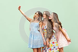 Beautiful three young adult fasion model, macking selfie, grimacing and tongue out. Green background.