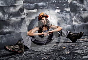 Beautiful tired miner worker in orange helmet with flashlight sitting on a floor on steel background