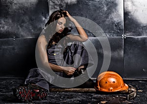 Beautiful tired miner worker sitting on a floor on steel background, pickaxe and orange helmet lies nearby