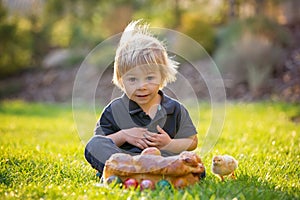Beautiful toddler boy, eating sweet bread and eggs in garden on sunset, little chicks running around