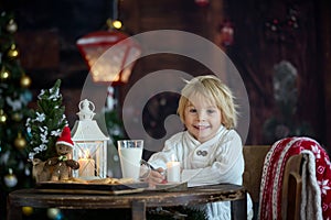 Beautiful toddler child, blond boy, writing letter to Santa Claus and eating cookies at home, cozy atmosphere