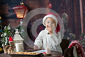 Beautiful toddler child, blond boy, writing letter to Santa Claus and eating cookies at home, cozy atmosphere