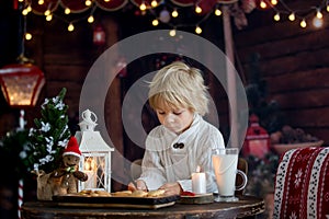 Beautiful toddler child, blond boy, writing letter to Santa Claus and eating cookies at home, cozy atmosphere