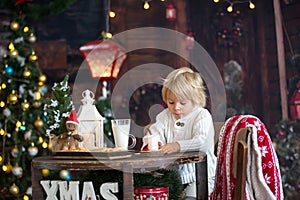 Beautiful toddler child, blond boy, writing letter to Santa Claus and eating cookies at home, cozy atmosphere