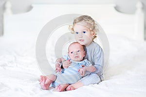 Beautiful toddler girl holding her newborn baby brother on a white bed