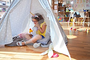 Beautiful toddler wearing glasses and unicorn diadem sitting on the floor at kindergarten shocked covering mouth with hands for