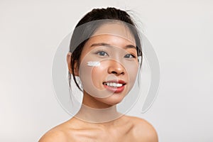 Beautiful topless young korean lady with moisturizer cream on her face, testing new beauty product, white background