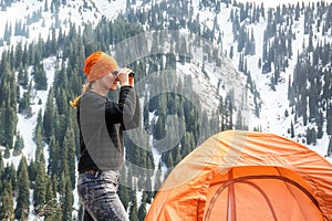 Beautiful tourist girl standing near a tent and looking through binoculars at snow-capped mountains