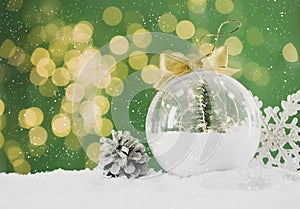 Beautiful transparent Christmas ornament with small fir tree and fairy lights on snow against green background, bokeh effect.