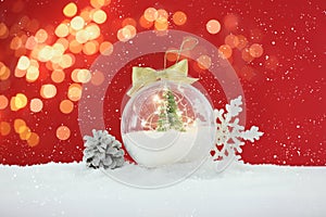 Beautiful transparent Christmas ornament with small fir tree and fairy lights on snow against red background, bokeh effect