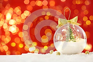 Beautiful transparent Christmas ornament with small fir tree and fairy lights on snow against red background, bokeh effect. Space
