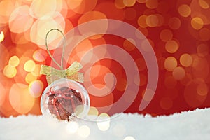 Beautiful transparent Christmas ornament with small gift box on snow against red background, bokeh effect. Space for text