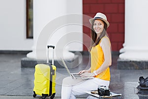 Beautiful traveler tourist woman in hat with suitcase sit using work on laptop pc computer looking aside in city outdoor