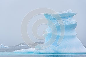 Beautiful turquoise blue iceberg floating in the Antarctic, against a foggy background with a research station