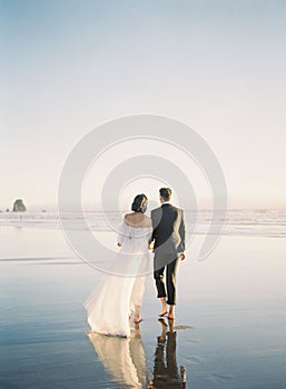 Beautiful vertical back shot of a newly wed couple walking barefoot on the beach