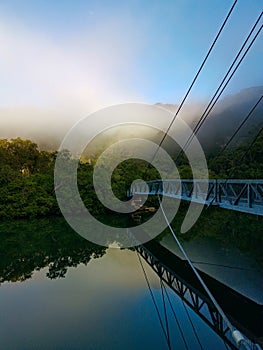 Beautiful view of creek with reflections suspension bridge, blue sky, light clouds, fog, mountains and trees on water