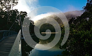 Beautiful view of creek with reflections suspension bridge, blue sky, light clouds, fog, mountains and trees on water