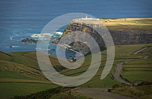 Beautiful view of green fields and single lighthouse standing on steep brink of sea coast. Bright blue water and rocks