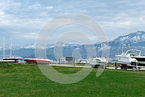 Beautiful view of green lawn in front of the embankment with boats and yacht on land on trailer against the backdrop of snow-c