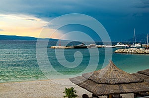 Beautiful view of sandy beach with straw umbrellas, port and small lighthouse on stone pier in front of Brac island on the eve of