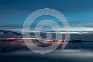 Beautiful view of Trasimeno lake Umbria, Italy at dusk, with blue and orange tones and moon in the sky