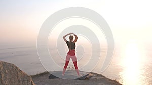 Beautiful view of woman doing yoga stretching on the mountain with sea view at sunset. Stretching arms.