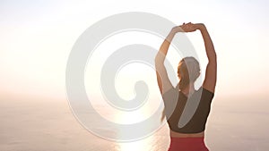 Beautiful view of woman doing yoga stretching on the mountain with sea view at sunset. Stretching hands up. Back view.
