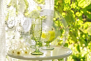 Beautiful vintage glass of white wine and sweets with white flowers on glass table in summer garden in sunlight.