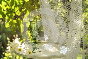 Beautiful vintage glass of white wine with white flowers on glass table in summer garden in sunlight.