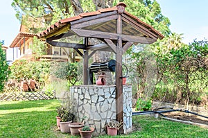 Beautiful Vintage Water Well with Wooden Roof and Tiles, Decorated with Plants and Flower Pots in a Backyard with Grass and Trees