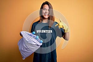 Beautiful volunteer girl caring environment doing volunteering holding bag with rubish bottles with surprise face pointing finger