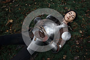 A beautiful warrior girl wearing chainmail and armor lying on the ground in a mysterious forest.