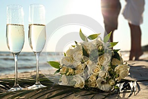 Beautiful wedding bouquet, glasses of champagne and couple on sandy beach