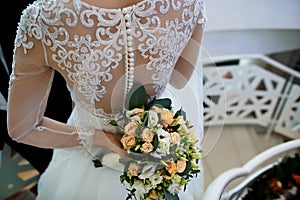 Beautiful wedding bouquet with white flowers and green leaves in the hands of the bride and groom in a dress with a lace back