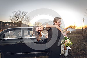 Beautiful wedding couple in the countryside next to the retro car. man groom is taking holding bride in his arms. smiling happy ch