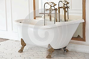 A beautiful white Baroque bathtub with gold legs and a faucet stands in a bright, spacious room next to a large mirror.