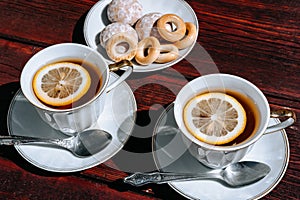 Beautiful white ceramic tableware for tea drinking. Two white cups of black tea with lemon on a wooden table, and delicious bagels