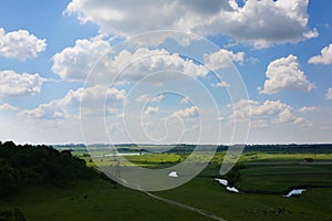 Beautiful white clouds in the blue sky. Amazing summer view on the field with shades. Green lawn and river with houses in the