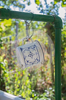 Beautiful white metal mug with blue decorations hanging on the pipes of outdoor sink in the garden