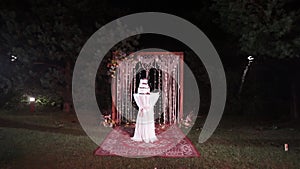 A beautiful white original handmade wedding cake stands in the lighting near the wedding arch in the evening