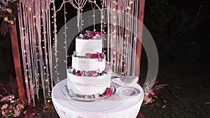 A beautiful white original handmade wedding cake stands in the lighting near the wedding arch in the evening. camera