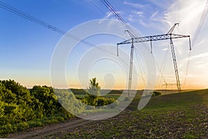 Beautiful wide panorama of high voltage lines and power pylons stretching through spring fields above group of green trees at dawn