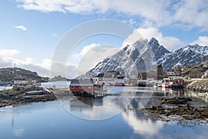 Beautiful winter fishing town on Nordic shore with tall snow mountain backdrop and reflection on the water surface.