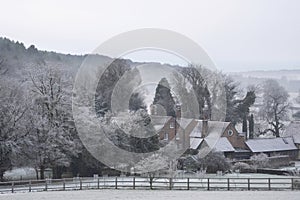 Beautiful Winter landscape of farm in fog in English countryside at dawn with hoarfrost covering trees and buildings