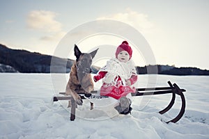 Beautiful winter. Little baby girl with her best friend dog sit on old wooden sledge in snow-covered landscape. Children with pet