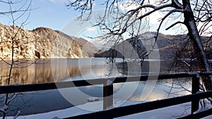 Beautiful winter mountain lake. Railings and trees in front of water and snowy mounts.