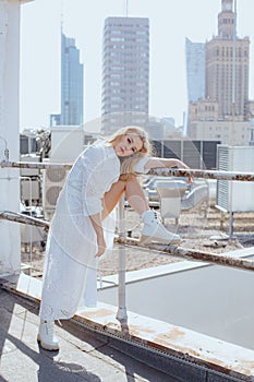 Beautiful woman against background of urban architecture. Charming blonde in snow-white dress poses on city roo.