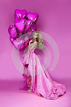Beautiful woman in amazing long ball dress is holding pink balloons and is posing on pink background. Elegant female model in gown