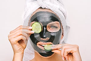 Beautiful woman applying black purifying charcoal facial mask with cucumber slices. Face skin care